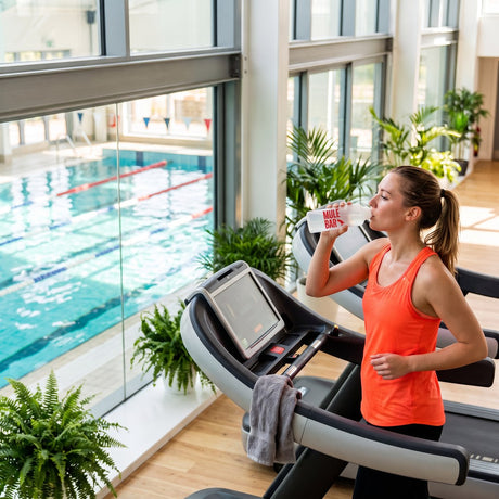 Coureuse sur tapis de course qui boit à son bidon Mulebar, piscine intérieure en arrière-plan, dans une salle baignée de lumière